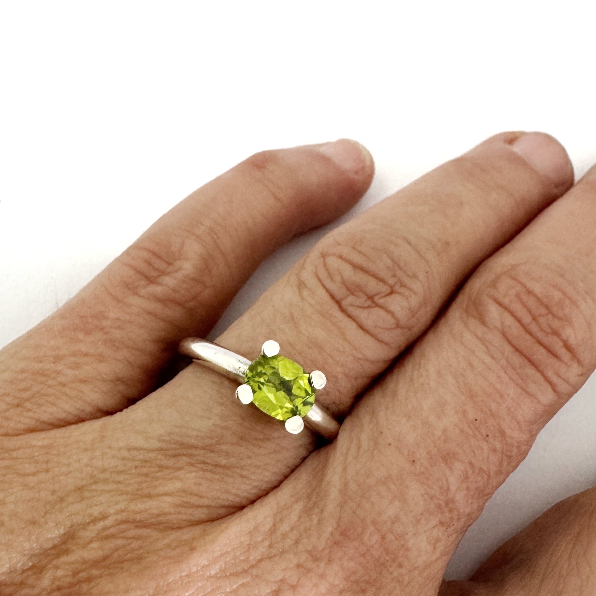 A silver ring with a oval green peridot gemstone worn on a person's finger, shown against a plain white background.
